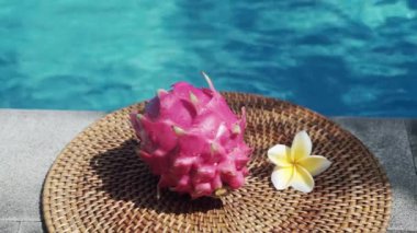 Video görüntüleri, taze kırmızı ejderha meyvesiyle yüzme havuzunun kenarında beyaz tropikal çiçeklerle frangipani 'yi, palmiye dalından gölgeyi ve Bali' de arka planda kaynayan mavi havuz suyunu gösteriyor. 