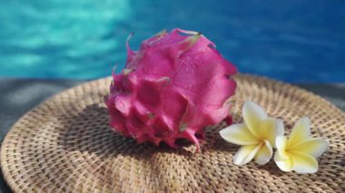 Video görüntüleri, taze kırmızı ejderha meyvesiyle yüzme havuzunun kenarında beyaz tropikal çiçeklerle frangipani 'yi, palmiye dalından gölgeyi ve Bali' de arka planda kaynayan mavi havuz suyunu gösteriyor. 
