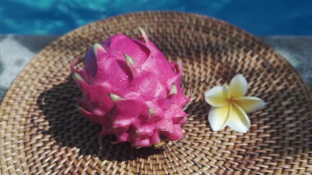 ビデオ映像は 白い熱帯の花のフランジパニを持つスイミングプールの端に澄んだ水の滴と新鮮な赤いドラゴンフルーツの終わり ヤシの枝から日陰を移動し バリ島の背景に青い水を泡立てるプール — ストック動画