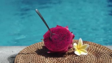 Taze kırmızı ejderha meyvesi, bir kaşıkla iki parçaya bölünmüş, köpüren mavi yüzme havuzunun kenarında, beyaz tropikal çiçekler, frangipani, Bali 'deki palmiye dalından gelen gölgeyi hareket ettiriyor.