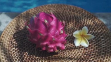 Video görüntüleri, taze kırmızı ejderha meyvesiyle yüzme havuzunun kenarında beyaz tropikal çiçeklerle frangipani 'yi, palmiye dalından gölgeyi ve Bali' de arka planda kaynayan mavi havuz suyunu gösteriyor. 