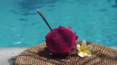 Taze kırmızı ejderha meyvesi, bir kaşıkla iki parçaya bölünmüş, köpüren mavi yüzme havuzunun kenarında, beyaz tropikal çiçekler, frangipani, Bali 'deki palmiye dalından gelen gölgeyi hareket ettiriyor.