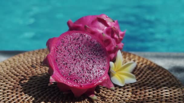 新鲜的红色龙果被切割成两半 新鲜的果肉在冒着泡沫的蓝色游泳池边 白色的热带花 从巴厘的棕榈枝移出阴影 — 图库视频影像