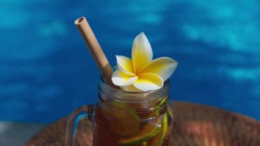 Buzlu çay ile cam mason kavanozunun video görüntüleri, buz küpleri ve limon dilimleri ile soğuk bambu kamışı, tam limon, frangipani çiçeği ve arka planda kaynayan mavi yüzme havuzu..