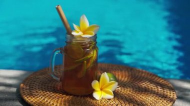 Buzlu çay ile cam mason kavanozunun video görüntüleri, buz küpleri ve limon dilimleri ile soğuk bambu kamışı, tam limon, frangipani çiçeği ve arka planda kaynayan mavi yüzme havuzu..