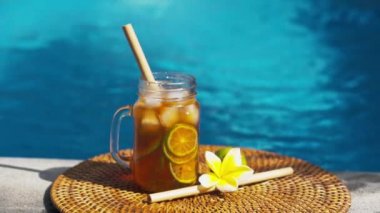Buzlu çay ile cam mason kavanozunun video görüntüleri, buz küpleri ve limon dilimleri ile soğuk bambu kamışı, tam limon, frangipani çiçeği ve arka planda kaynayan mavi yüzme havuzu..