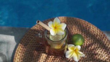 Buzlu çay ile cam mason kavanozunun video görüntüleri, buz küpleri ve limon dilimleri ile soğuk bambu kamışı, tam limon, frangipani çiçeği ve arka planda kaynayan mavi yüzme havuzu..