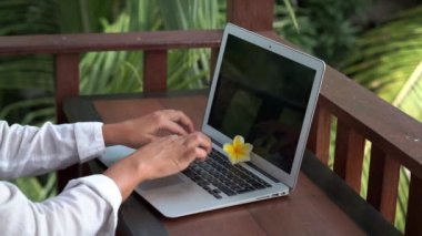 Genç sarışın kadının dizüstü bilgisayarla çalıştığı video görüntüleri, Bali 'de palmiye ağaçları manzaralı tropikal bungalovun balkonunda bikini ve beyaz gömlek yazarken.