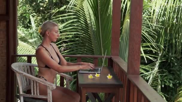 若いブロンドの女性のビデオ映像ラップトップで作業フリーランス バリ島でヤシの木のビューとトロピカルバンガローのバルコニーにビキニを入力 — ストック動画