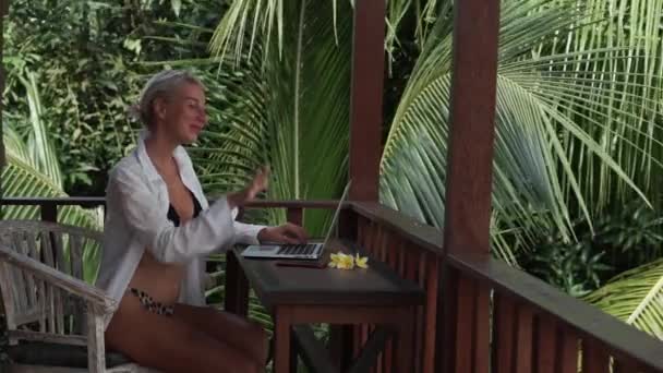 若いブロンドの女性のビデオ映像は ラップトップで作業フリーランス バリ島でヤシの木のビューとトロピカルバンガローのバルコニーにビキニと白いシャツを入力 — ストック動画