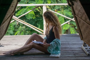 Kıvırcık sarı saçlı genç kadın Bali 'deki tropik bungalovun balkonunda kitap okuyor.