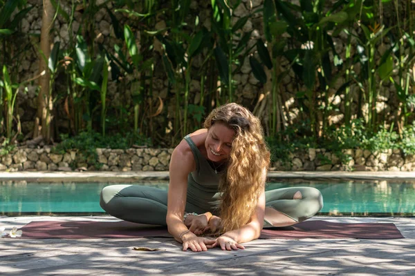 在巴厘岛 坐在蓝色游泳池和花园旁边的年轻女子穿着卡纳萨纳瑜伽 一头卷曲的金发 蓝眼睛 — 图库照片