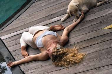Uzun kıvırcık sarı saçlı, yoga yapan mavi gözlü ve Bali 'deki mavi yüzme havuzunun yanında beyaz köpekli genç bir kadın..