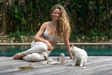 Sarı saçlı, beyaz tozlu, beyaz kedili, mavi yüzme havuzunun yanında büyük kristaller ve Bali 'deki bahçeli mavi gözlü genç bir kadın..