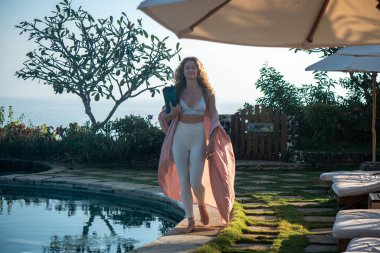 Kıvırcık sarı saçlı, beyaz taytlı, mavi gözlü, elinde yoga minderiyle yüzme havuzunun kenarında yüzen, Bali 'de okyanus manzaralı genç bir kadın.