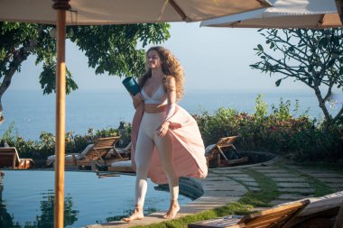 Kıvırcık sarı saçlı, beyaz taytlı, mavi gözlü, elinde yoga minderiyle yüzme havuzunun kenarında yüzen, Bali 'de okyanus manzaralı genç bir kadın.