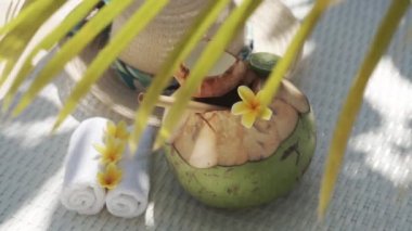 Video footage of green young coconut close up with bamboo straw, sun hat, tropical flowers frangipani, palm shade on white sun bed in Bali 