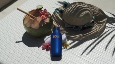 Video footage of green young coconut close up with bamboo straw, bottle of sunscreen SPF 50, sun hat, tropical pink flowers frangipani, palm shade on white sun bed in Bali 