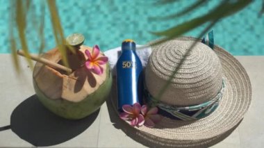 Video footage of green young coconut close up with bamboo straw, bottle of sunscreen SPF 50, sun hat, tropical pink flowers frangipani, palm shade on the edge of swimming pool in Bali