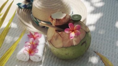 Video footage of green young coconut close up with bamboo straw, sun hat, tropical flowers frangipani, palm shade on white sun bed in Bali 