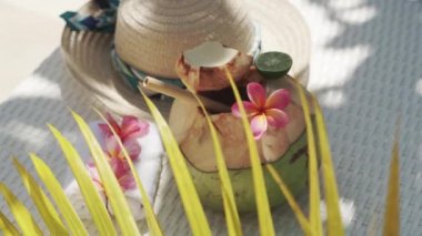 Video footage of green young coconut close up with bamboo straw, sun hat, tropical flowers frangipani, palm shade on white sun bed in Bali 