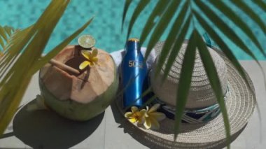 Yeşil hindistancevizinin video görüntüleri bambu kamışı, bir şişe güneş kremi 50, güneş şapkası, tropik sarı çiçek frangipani, Bali 'de yüzme havuzunun kenarında palmiye gölgesi