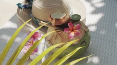 Video footage of green young coconut close up with bamboo straw, sun hat, tropical flowers frangipani, palm shade on white sun bed in Bali 