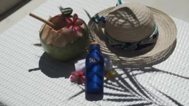 Video footage of green young coconut close up with bamboo straw, bottle of sunscreen SPF 50, sun hat, tropical pink flowers frangipani, palm shade on white sun bed in Bali 