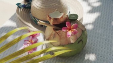 Video footage of green young coconut close up with bamboo straw, sun hat, tropical flowers frangipani, palm shade on white sun bed in Bali 