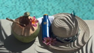 Video footage of green young coconut close up with bamboo straw, bottle of sunscreen SPF 50, sun hat, tropical pink flowers frangipani, palm shade on the edge of swimming pool in Bali