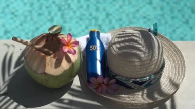 Video footage of green young coconut close up with bamboo straw, bottle of sunscreen SPF 50, sun hat, tropical pink flowers frangipani, palm shade on the edge of swimming pool in Bali