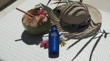Video footage of green young coconut close up with bamboo straw, bottle of sunscreen SPF 50, sun hat, tropical pink flowers frangipani, palm shade on white sun bed in Bali 