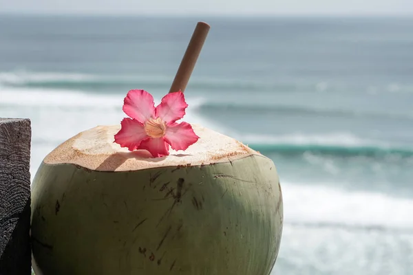 緑の若い新鮮なココナッツの近くに竹のわらとピンクの熱帯の花が崖と青い海にあり 大きな波がバリ島でサーフィンをしています — ストック写真