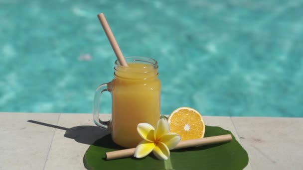 オレンジジュース 新鮮なオレンジの半分 黄色Frangipani花と背景にバブルブルースイミングプールとガラスのメイソンジャーのビデオ映像 — ストック動画