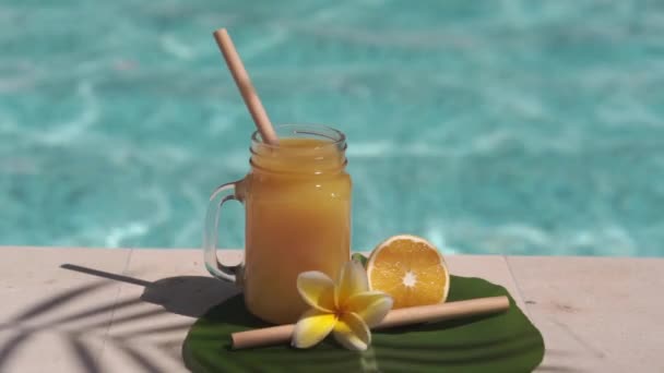 有橙汁 一半新鲜橙子 一半黄色乳酪花 棕榈树树荫和背景上起泡的蓝色游泳池的玻璃瓶视频镜头 — 图库视频影像