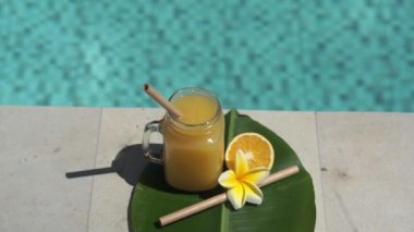 Portakal suyu, bambu kamışı, taze portakal, sarı frangipani çiçeği ve arka planda kaynayan mavi yüzme havuzunun olduğu cam mason kavanozunun video görüntüleri..