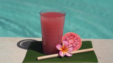 Pembe guava suyu, bambu kamışı, taze pembe guava, tropikal çiçek frangipani ve arka planda kaynayan mavi yüzme havuzunun video görüntüleri..