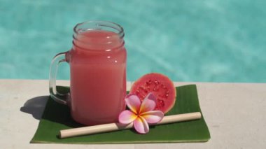 Cam mason kavanozu, pembe guava suyu, bambu kamışı, taze pembe guava, tropikal çiçek frangipani ve arka planda kaynayan mavi yüzme havuzunun video görüntüleri..