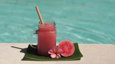 Cam mason kavanozu, pembe guava suyu, bambu kamışı, taze pembe guava, tropikal çiçek frangipani ve arka planda kaynayan mavi yüzme havuzunun video görüntüleri..