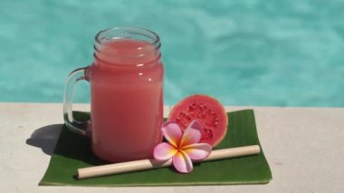 Cam mason kavanozu, pembe guava suyu, bambu kamışı, taze pembe guava, tropikal çiçek frangipani ve arka planda kaynayan mavi yüzme havuzunun video görüntüleri..