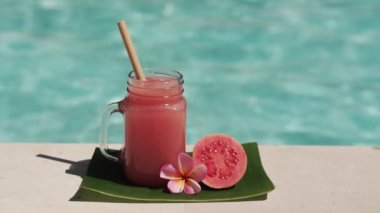 Cam mason kavanozu, pembe guava suyu, bambu kamışı, taze pembe guava, tropikal çiçek frangipani ve arka planda kaynayan mavi yüzme havuzunun video görüntüleri..