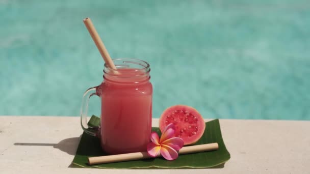 ピンク色のグアバジュース 竹の藁 新鮮なピンク色のグアバの半分 熱帯の花Frangipaniと背景にバブルブルースイミングプールとガラスのマソン瓶のビデオ映像 — ストック動画