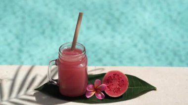 Pembe guava suyu, bambu kamışı, taze pembe guava, tropik çiçek frangipani, palmiye ağacının gölgesi ve arka planda kaynayan mavi yüzme havuzu olan cam mason kavanozunun video görüntüleri..