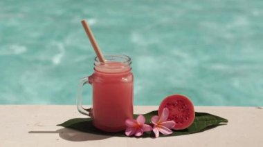Cam mason kavanozu, pembe guava suyu, bambu kamışı, taze pembe guava, tropikal çiçek frangipani ve arka planda kaynayan mavi yüzme havuzunun video görüntüleri..