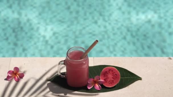 ピンクのグアバジュース 竹のわら 新鮮なピンクのグアバの半分 熱帯の花Frangipani ヤシの木から日陰 背景にバブルブルースイミングプールとガラスのマソン瓶のビデオ映像 — ストック動画