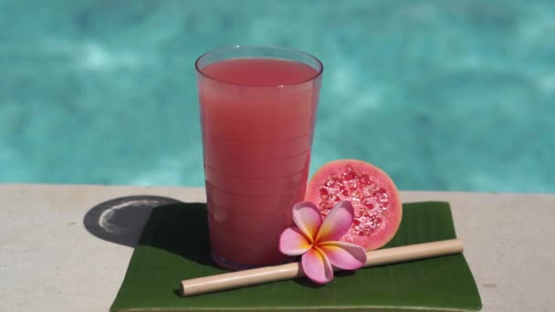 ピンク色のグアバジュース 竹の藁 新鮮なピンク色のグアバの半分 熱帯の花Frangipaniと背景にバブルブルースイミングプールとガラスのビデオ映像 — ストック動画