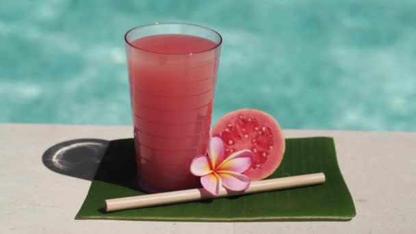 ピンク色のグアバジュース 竹の藁 新鮮なピンク色のグアバの半分 熱帯の花Frangipaniと背景にバブルブルースイミングプールとガラスのビデオ映像 — ストック動画