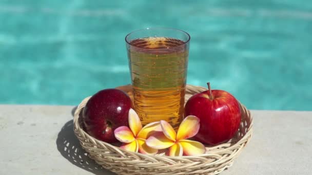 有苹果汁 红苹果 热带花 香草和泡沫蓝色游泳池背景的玻璃杯录像 — 图库视频影像