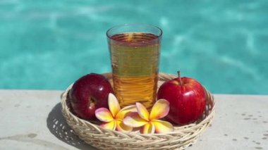 Elma suyu, bambu kamışı, kırmızı elma, tropikal çiçek frangipani ve arka planda kaynayan mavi yüzme havuzunun video görüntüleri..