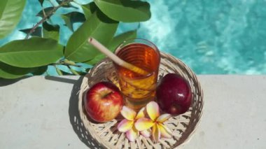 Elma suyu, bambu kamışı, kırmızı elma, tropikal çiçek frangipani, yeşil yapraklı ağaç dalı ve arka planda kaynayan mavi yüzme havuzu olan cam görüntüleri..